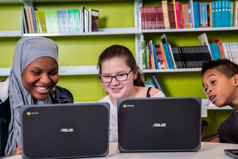 Three students sitting at a table and looking at one student's computer. 