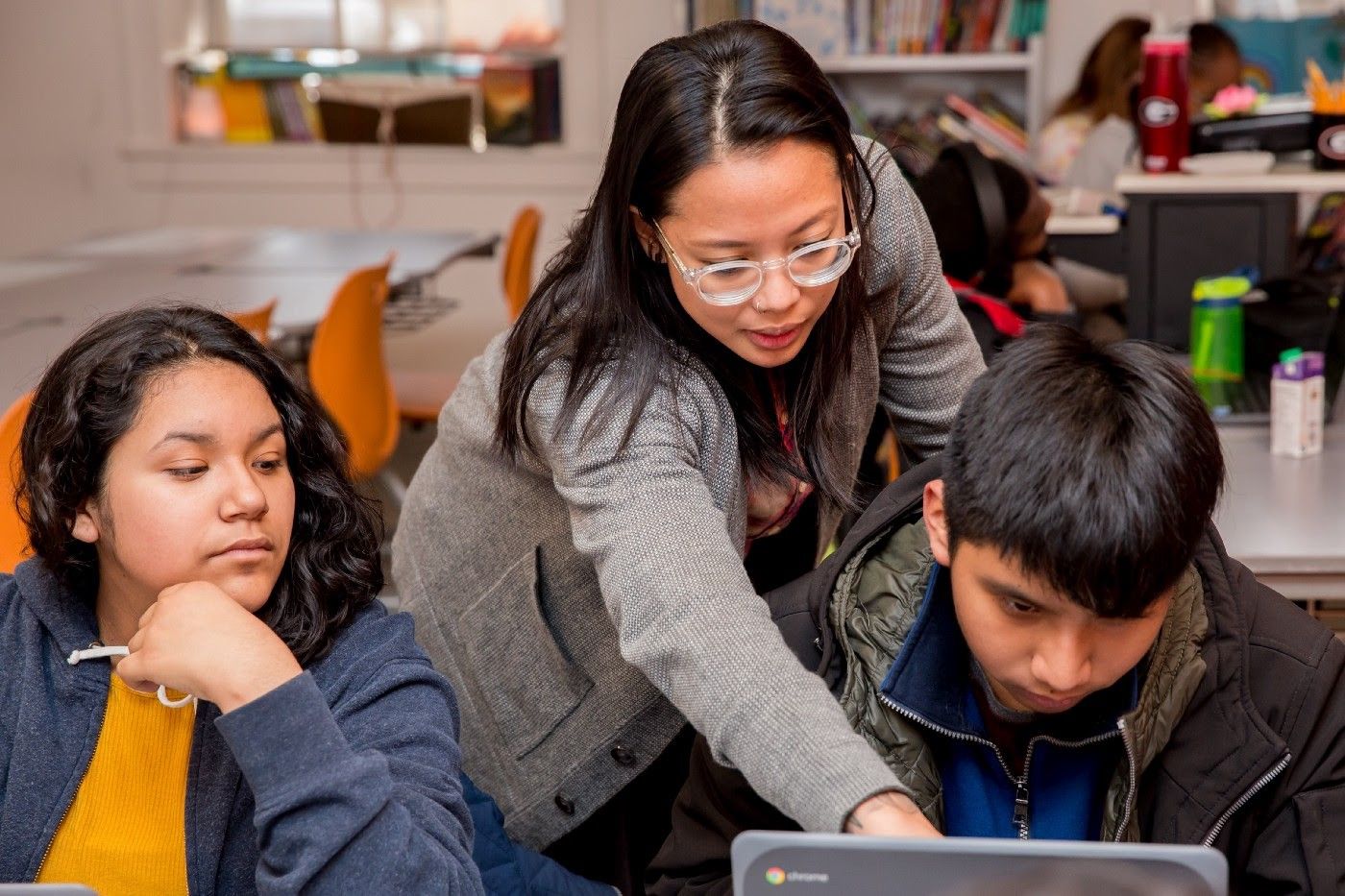 A teacher leaning over a student's shoulder and pointing at his computer screen. Another student sits next to them.