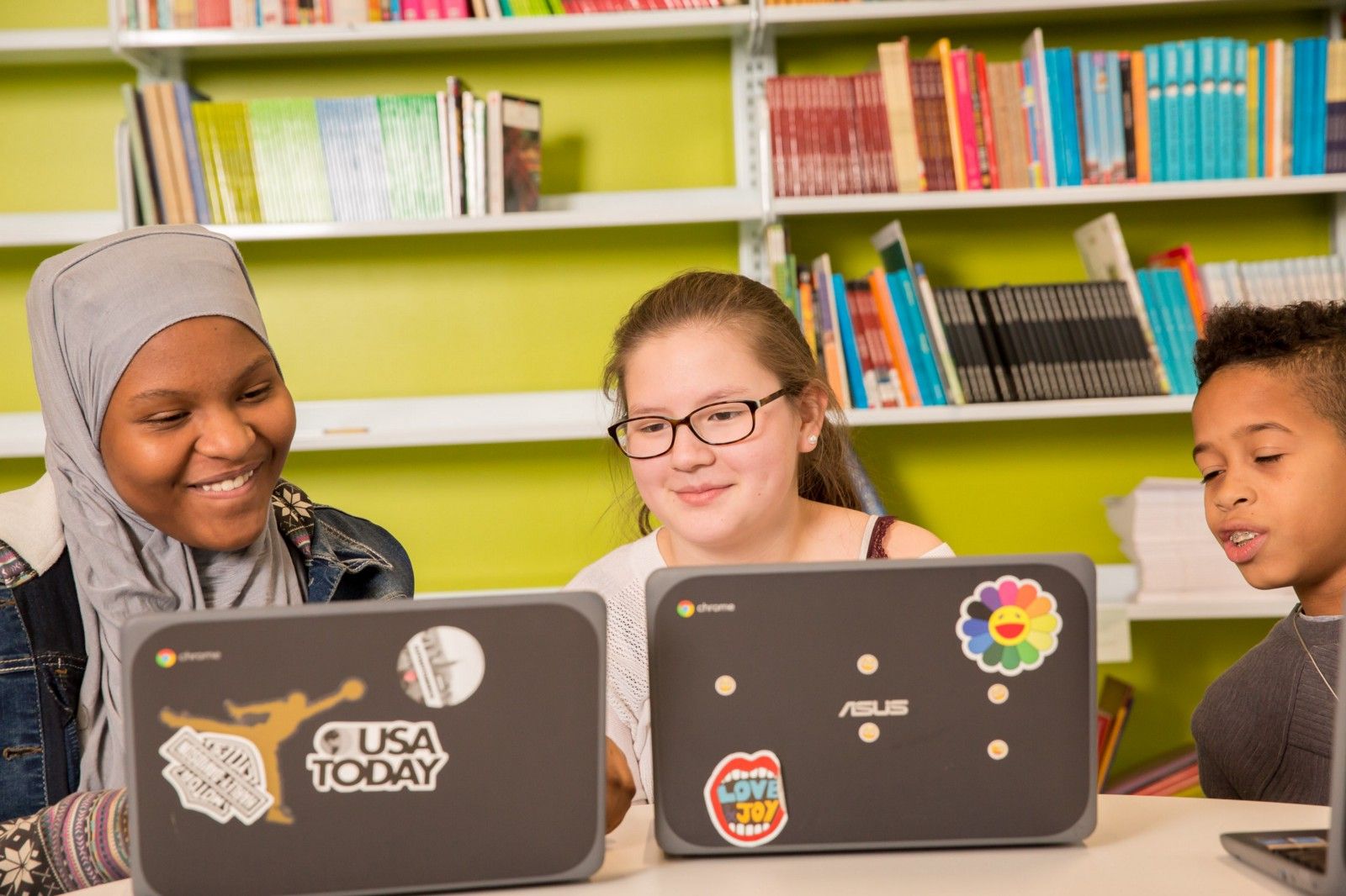 Three students sitting at a table and looking at one student's computer screen. 
