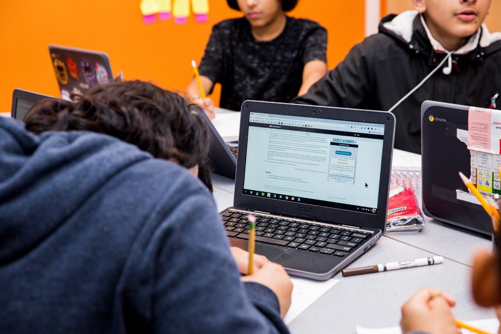 A group of students sitting at a table with their laptops open to a CommonLit lesson, writing on paper. 