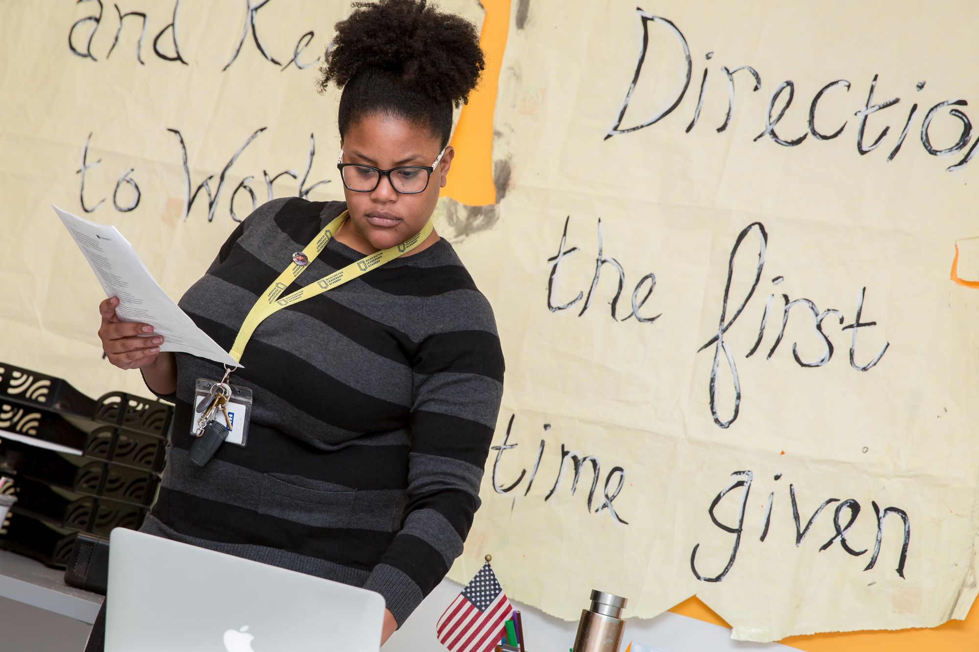 Teacher holds papers while typing on laptop.