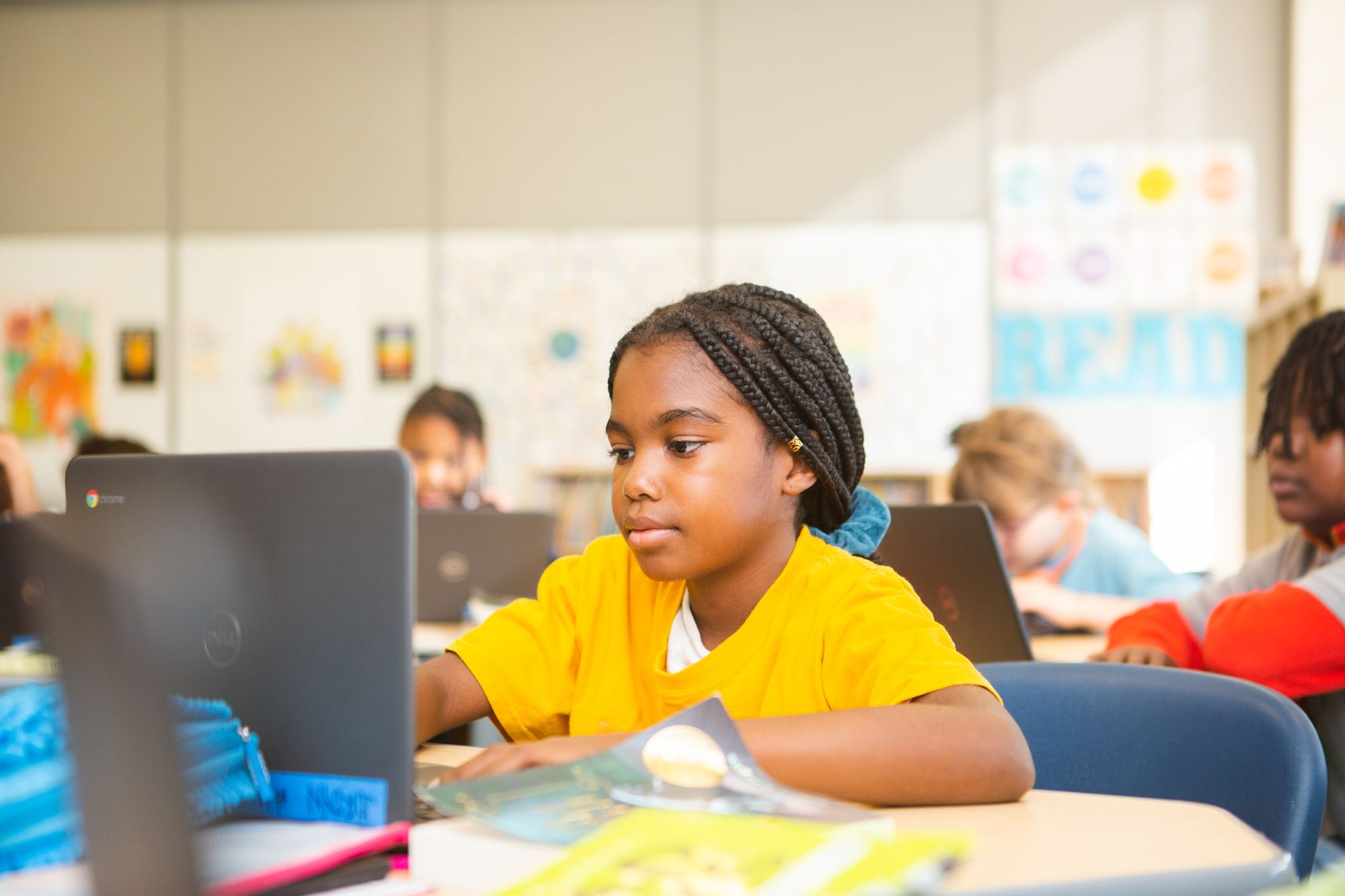 An elementary student works on a CommonLit Main Idea lesson. Other students in her class are visible in the background.