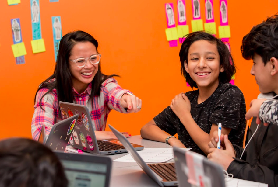 A teacher and two students sitting at a table. The teacher is pointing at a computer and smiling. 